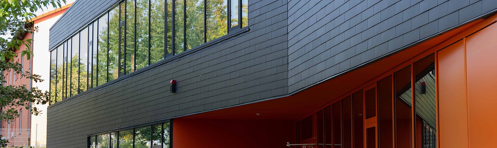 Fassade eines Gebäudes mit dunklem Schiefer und großen Fensterfronten. Der Eingangsbereich ist knallorange.
