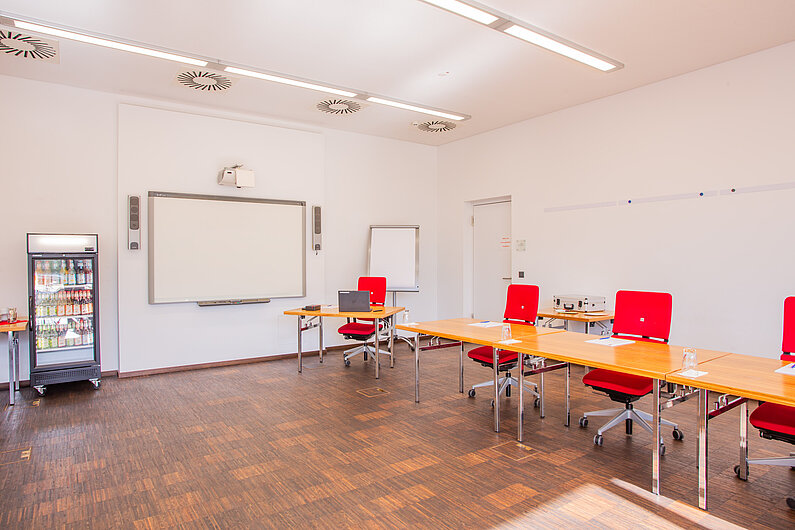 Raum mit Tischen und Drehstühlen daran. An der Wand ist ein Whiteboard, daneben steht ein Kühlschrank mit Getränken.
