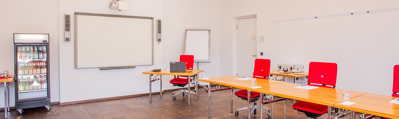 Raum mit Tischen und Drehstühlen daran. An der Wand ist ein Whiteboard, daneben steht ein Kühlschrank mit Getränken.